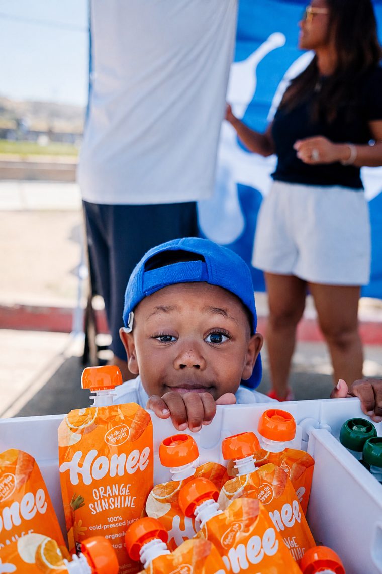 A toddler in a blue cap peeking over a cooler of Honee Orange Sunshine pouches
